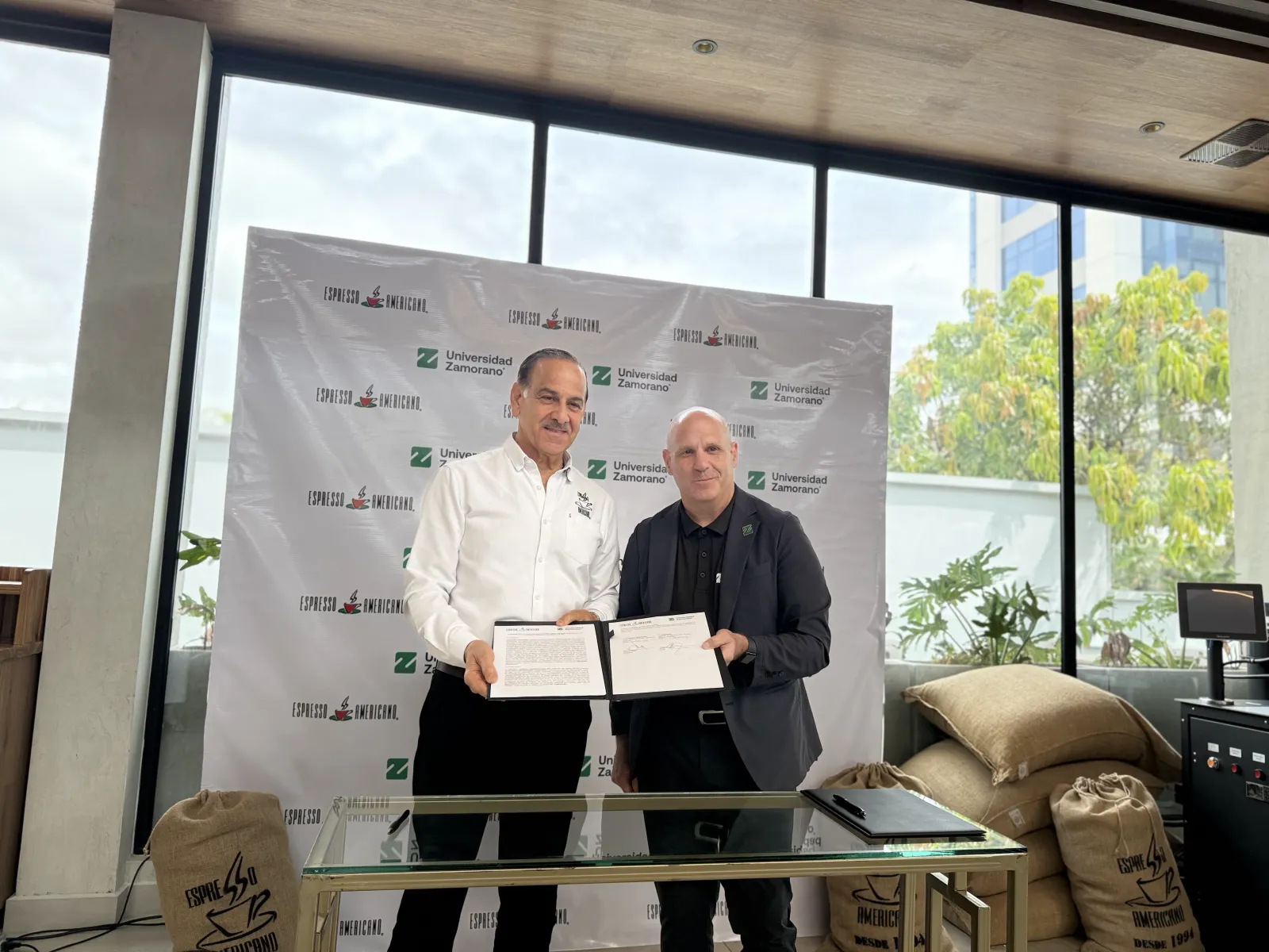 Representantes de Espresso Americano y Universidad Zamorano muestran el convenio firmado frente a un banner de ambas instituciones